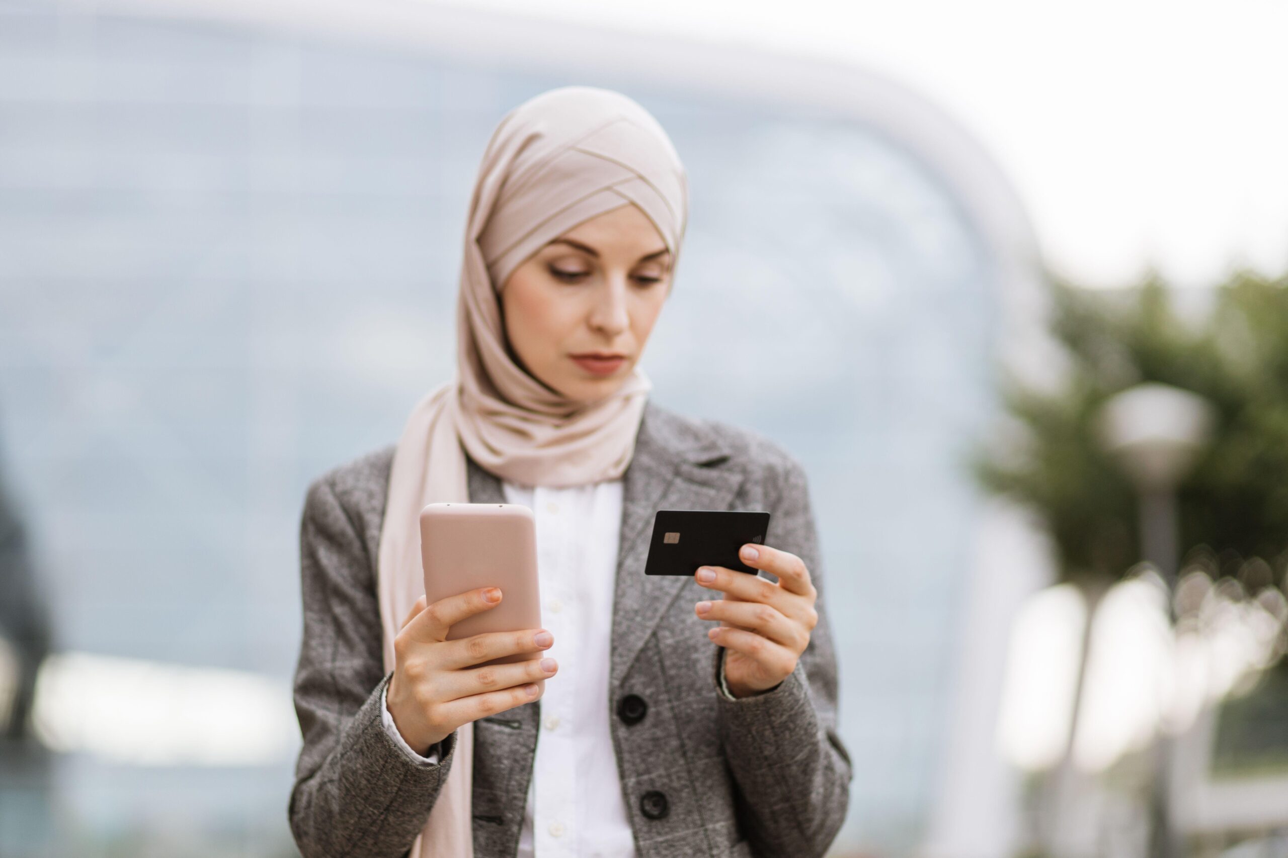 Pretty Muslim lady in hijab using smartphone and credit bank card for online shopping. Focused businesslady in headscarf standing on street and making purchase.