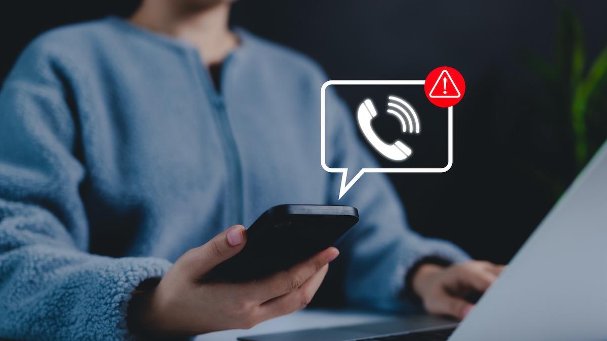 A woman's phone is buzzing with a scam as she looks at her laptop