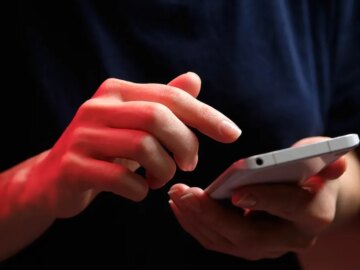 A mobile phone in the hands of a young woman in a dark colored t-shirt.