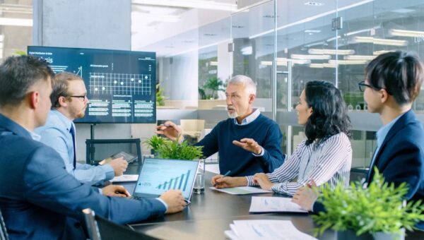 Senior Executive erklärt Vision und Potenzial des Unternehmens für seine Mitarbeiter. Sie sitzen am großen Tisch im Sitzungszimmer. Der Fernsehbildschirm an der Wand zeigt das Unternehmenswachstum.